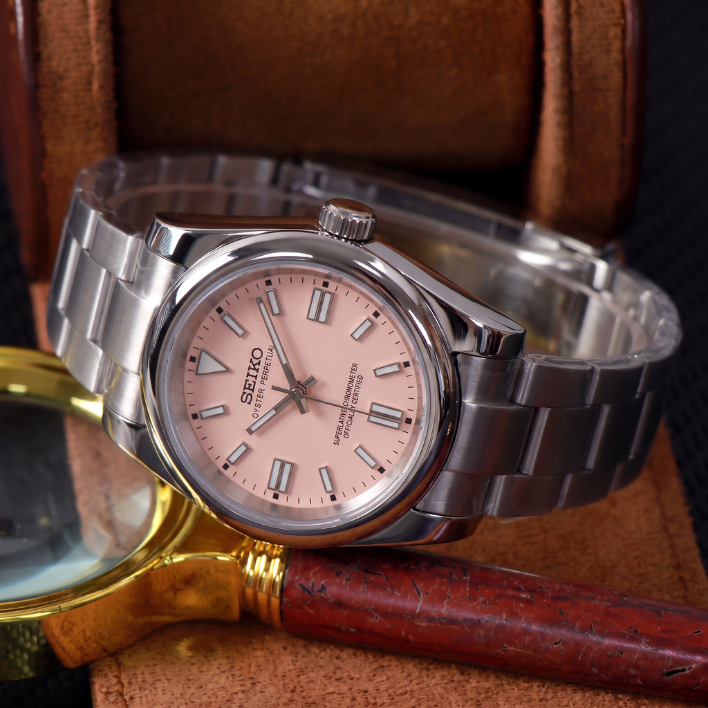 ORA Classic Sport-Style 36mm Seiko Mod watch, blush pink dial, smooth polished bezel, steel three-link bracelet – in a leather watch box beside a brass magnifying glass