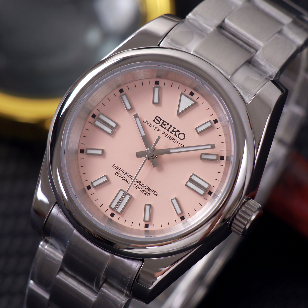 ORA Classic Sport-Style 36mm Seiko Mod watch, blush pink dial, smooth polished bezel, steel three-link bracelet – angled view against warm bokeh lights