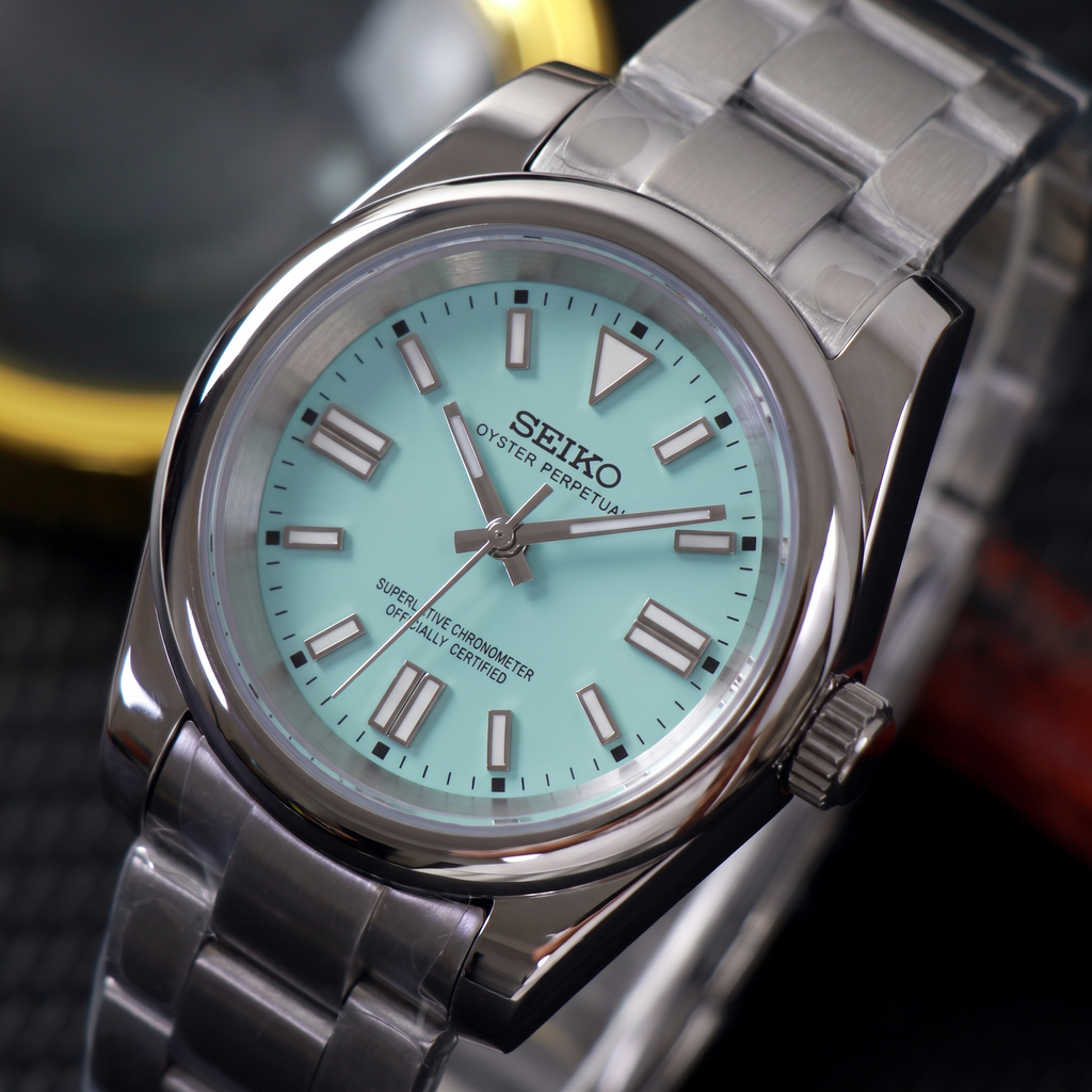 ORA Classic Sport-Style 36mm Seiko Mod watch, aqua dial, smooth polished bezel, steel three-link bracelet – angled view against warm bokeh lights